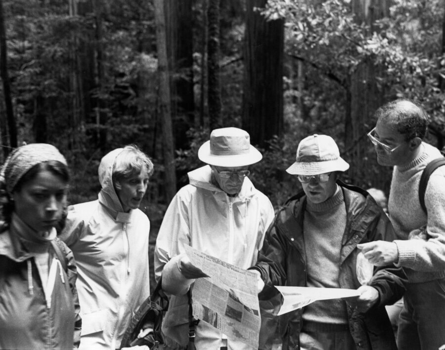 Five people stand together in a forest, wearing jackets and hats. Three of them are intently looking at maps or papers, possibly planning a hike or route, while the others observe the scene.
