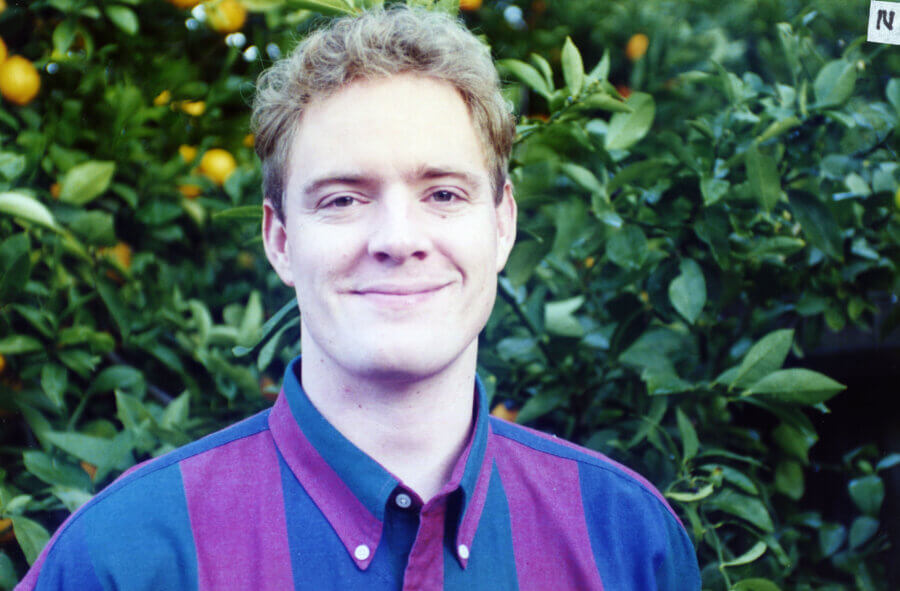 A person with short, light brown hair, wearing a maroon and blue striped collared shirt, smiles while standing in front of green leafy trees with yellow fruit.