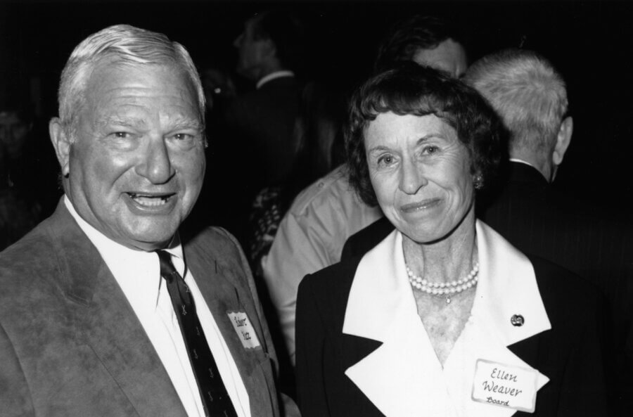 A person in a suit and a person in a blazer and pearls smile at the camera at a formal event. Both wear name tags. Other people are visible in the background. The image is black and white.
