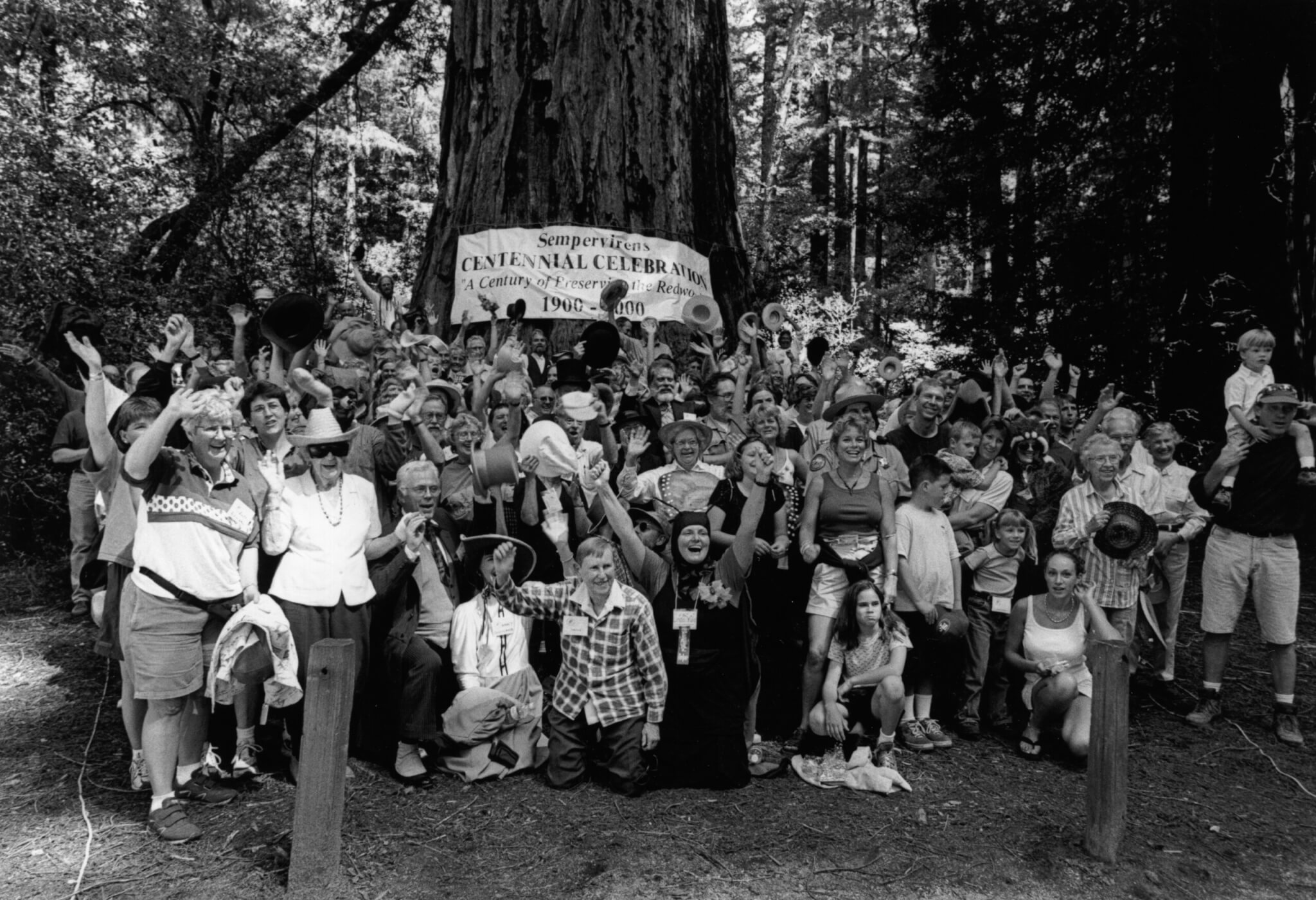 A large group of people wave for a photo in front of a giant tree, with a banner reading Sempervirens Centennial Celebration hanging behind them in a forested area.