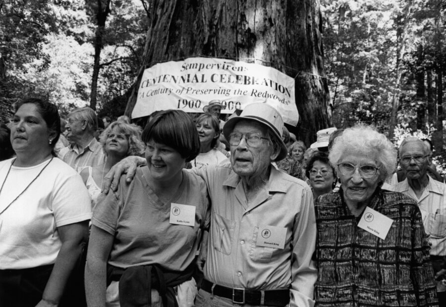 A group of people stand together outdoors in front of a large tree and a banner that reads “Sempervirens Centennial Celebration: A Century of Preserving the Redwoods 1900-2000.” Several people smile and pose for the photo.