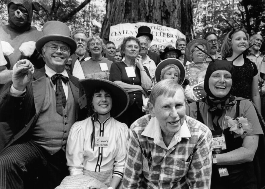 A group of people, some in vintage costumes and hats, pose and smile together outdoors in front of a large tree and a banner that reads “Sempervirens Centennial Celebration: A Century of Preserving the Redwoods 1900-2000.”