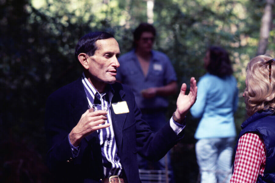 A person in a striped shirt and dark blazer gestures while talking to a person outdoors; three other people are in the background amid trees.