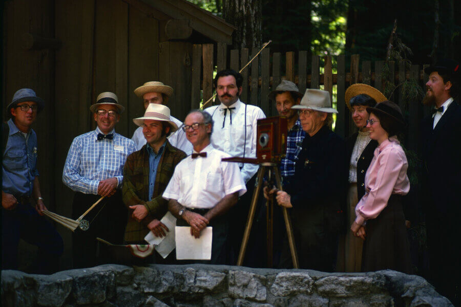 A group of people in vintage clothing poses outdoors by a wooden fence and building, gathered around an old-fashioned camera on a tripod. The setting appears to be historical or reenactment-themed.