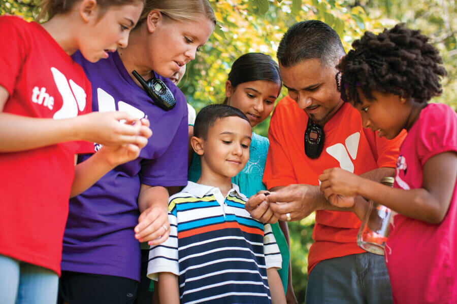 Two adults and four children stand closely together outdoors, examining a small object in one adult’s hand. The group looks curious and engaged, surrounded by greenery. Some wear shirts with a Y logo.