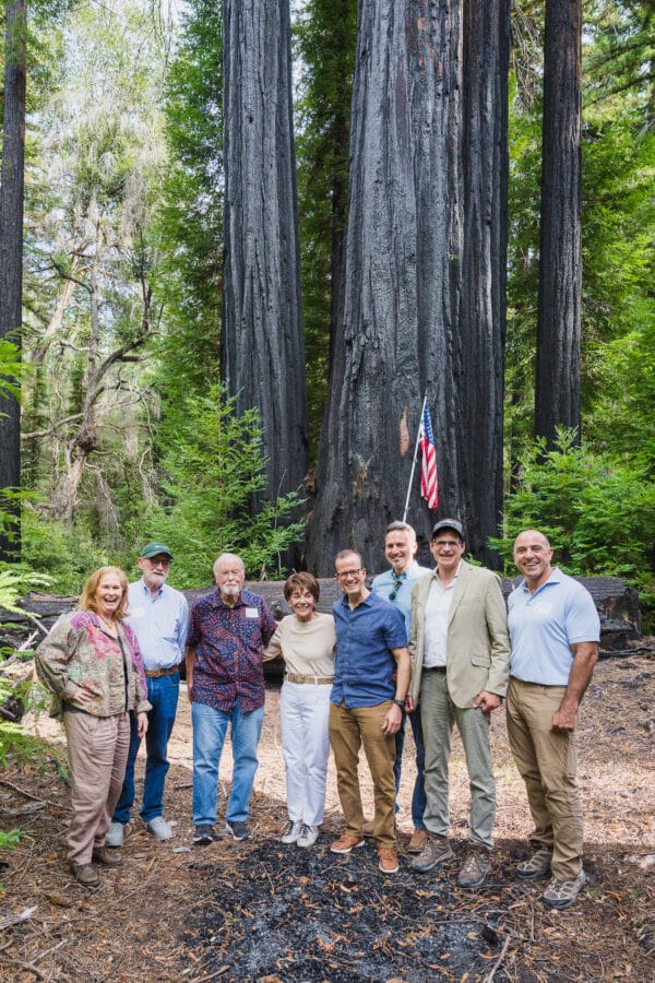 Eight people pose for a photo in Big Basin Redwoods State Park in front of a tree dedicated to Former Congresswoman Anna Eshoo. A United States Flag stands in front of the tree. (L-R) Assemblymember Gail Pellerin, State Senator John Laird, Former Congressman Sam Farr, Former Congresswoman Anna Eshoo, Assemblymember Marc Berman, Supervisor Ray Mueller, State Senator Josh Becker, and Congressman Jimmy Panetta; Photo credit: Orenda Randuch