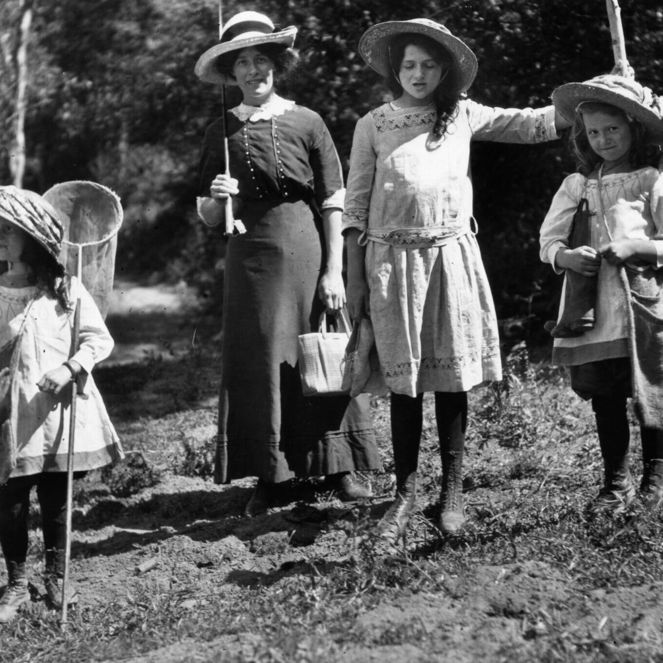 Four children in old-fashioned dresses and hats stand outdoors in a grassy, wooded area. Two hold fishing rods, one holds a net, and another has a small pail. The scene appears to be from the early 20th century.