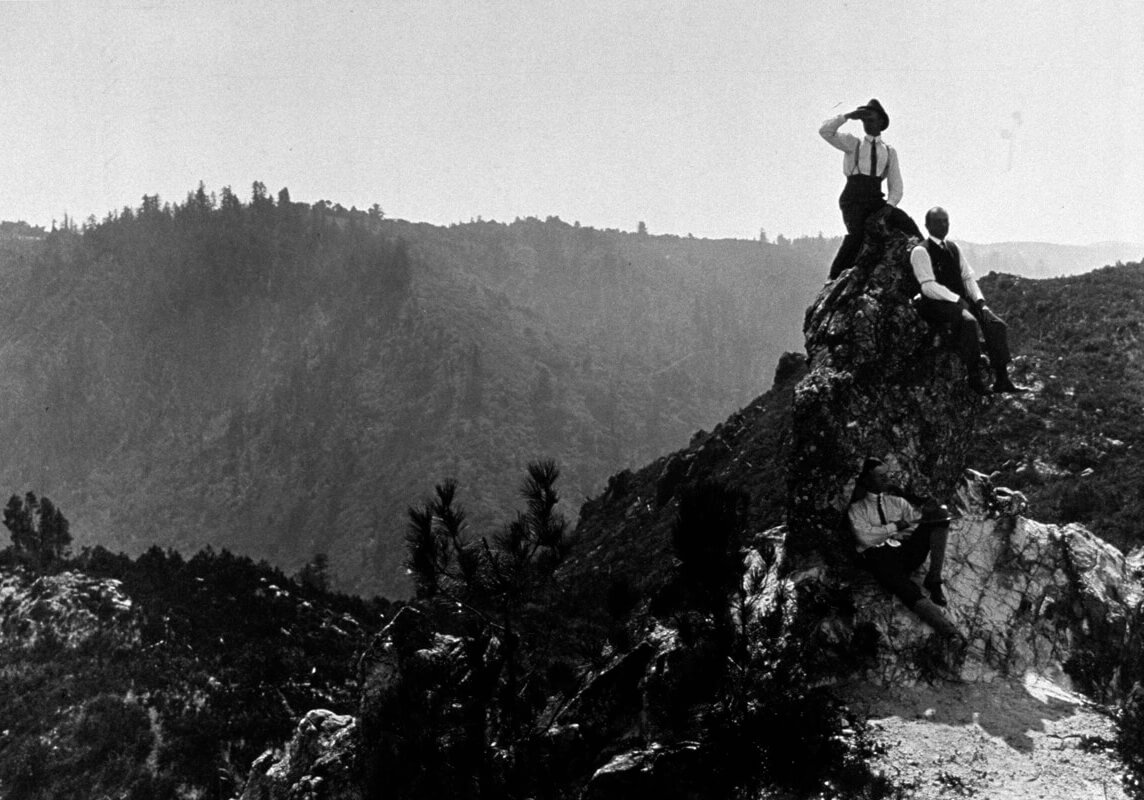 A black and white photo from Sempervirens Fund’s historic archive shows three men—in early 20th century suits consisting of dark colored trousers, white collared long-sleeved shirts with ties, dress shoes, and a few accessories like suspenders, vests, and hats reminiscent of fashion just after the turn of the century—sit casually atop a rock outcropping rising above the surrounding brush and forest like a spire as the view of forested mountainsides fade off into the distance behind them.